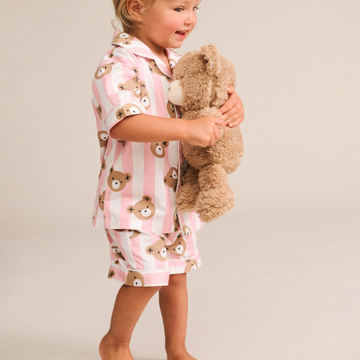 A toddler standing barefoot against a light beige studio background, wearing a pink and white striped pajama set with teddy bear face prints and holding a brown plush teddy bear.