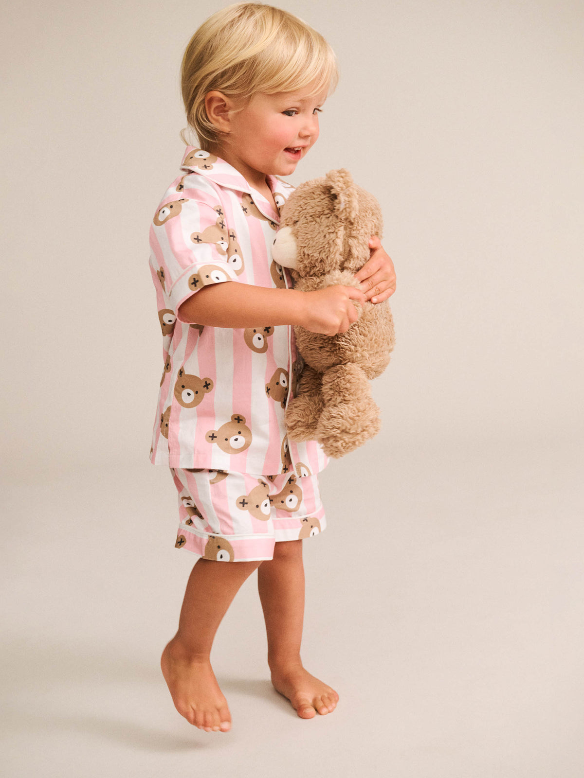 A toddler standing barefoot against a light beige studio background, wearing a pink and white striped pajama set with teddy bear face prints and holding a brown plush teddy bear.