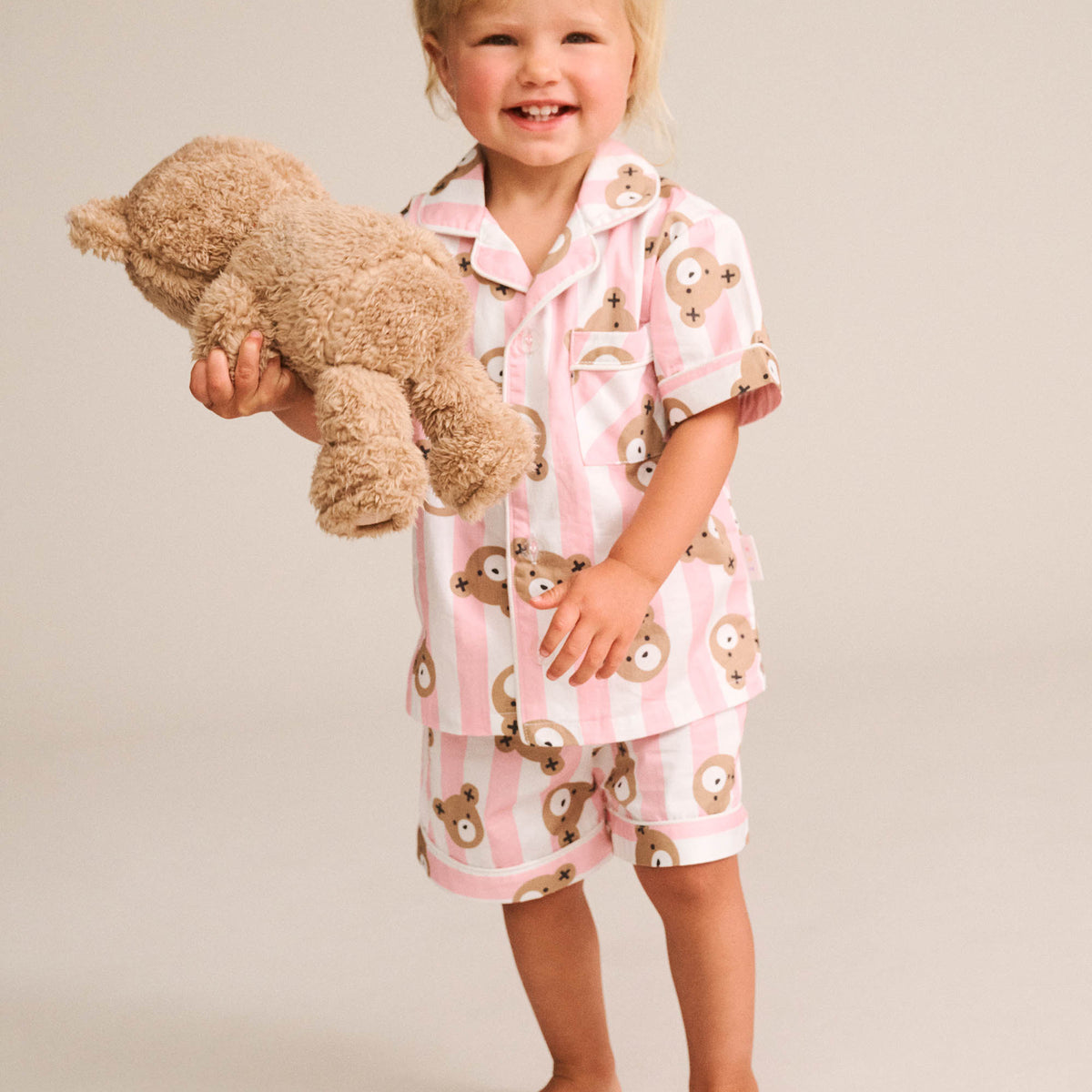 A smiling toddler stands barefoot against a plain light beige studio background, wearing a pink and white striped pajama set with a teddy bear face print and holding a plush teddy bear.