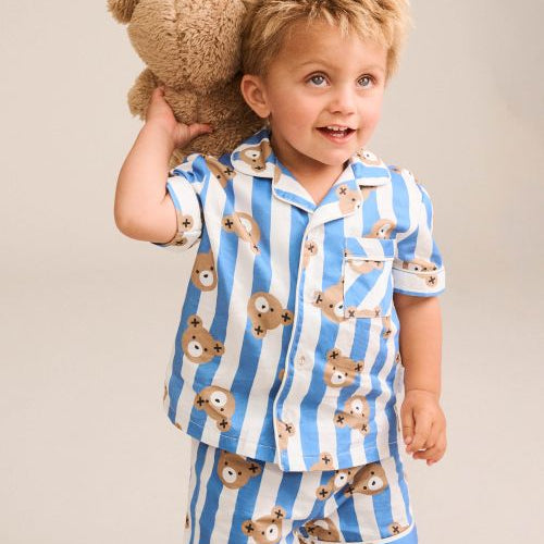 A young child wearing a blue and white striped pajama set with teddy bear prints, holding a brown teddy bear against their back, photographed against a solid light beige background.
