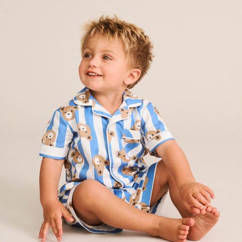 A young child sitting on a light beige studio floor wearing a blue and white striped short-sleeve pajama set with a teddy bear print.