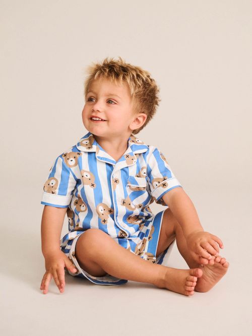 A young child sitting on a light beige studio floor wearing a blue and white striped short-sleeve pajama set with a teddy bear print.