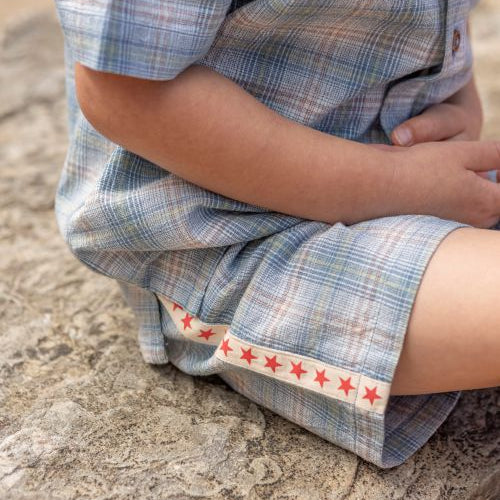 A close-up of a child sitting on a rock, wearing a light blue and white plaid short-sleeve shirt and shorts set with a decorative red star trim on the hem.