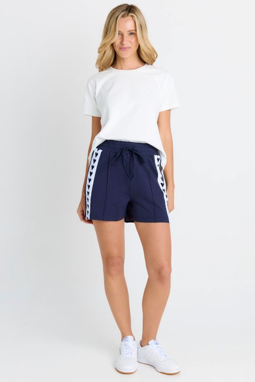 A woman modeling navy blue athletic shorts with white side stripes, paired with a white t-shirt and white sneakers, against a plain white studio background.