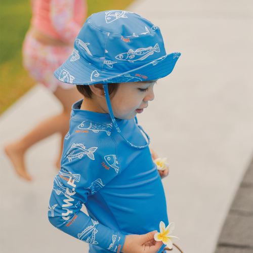 A toddler wearing a bright blue long-sleeve sun shirt and matching bucket hat with a white fish pattern, standing outdoors on a paved walkway.
