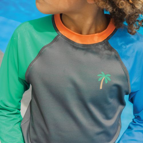 A close-up of a child wearing a long-sleeve rash guard with a gray body, green left sleeve, blue right sleeve, and an orange collar. A small palm tree logo is printed on the left chest.