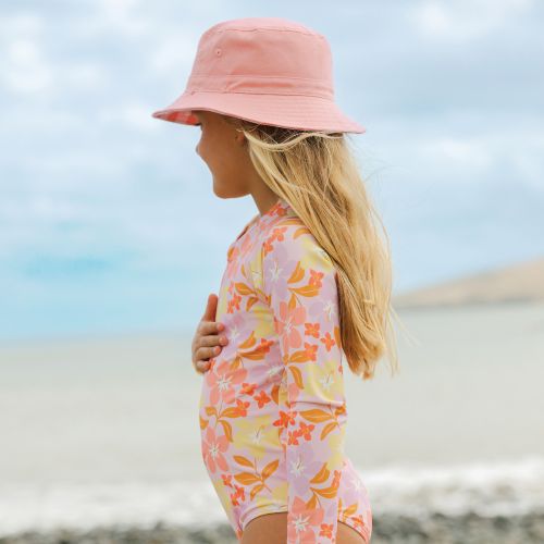 A young girl in profile on a beach, wearing a pink sun hat and a pink long-sleeve one-piece swimsuit with a yellow and orange floral pattern.