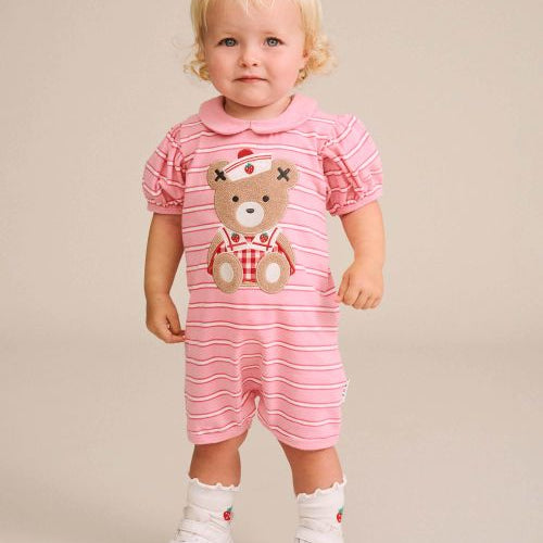 A toddler with blonde hair stands against a solid beige studio background wearing a pink and white striped short-sleeve romper with a teddy bear graphic on the front, paired with white socks and white sneakers.