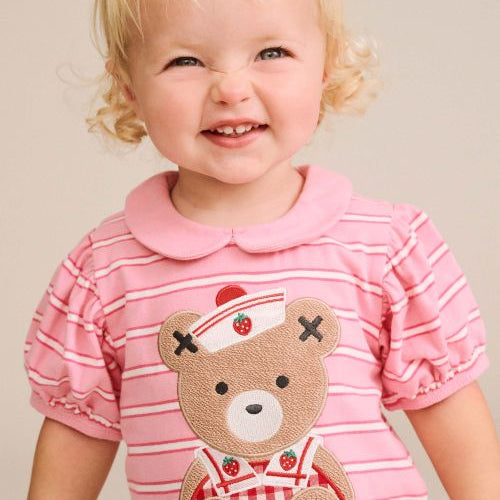 A smiling toddler with blonde hair wearing a pink short-sleeve striped shirt with a teddy bear applique on the front, photographed against a plain beige background.