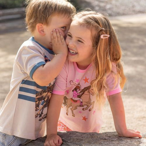 Two children sitting outdoors on a stone surface, with the girl wearing a pink short-sleeve shirt with a cowboy and horse graphic and the boy wearing a white graphic T-shirt. The background shows a blurred outdoor setting with trees and sunlight.
