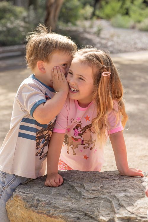 Two children sitting outdoors on a stone surface, with the girl wearing a pink short-sleeve shirt with a cowboy and horse graphic and the boy wearing a white graphic T-shirt. The background shows a blurred outdoor setting with trees and sunlight.