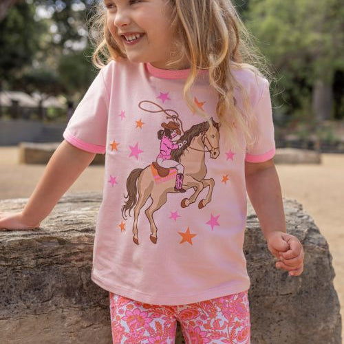 A young girl with blonde hair is smiling outdoors, wearing a light pink short-sleeve t-shirt with a cowboy on a horse graphic and pink patterned leggings.