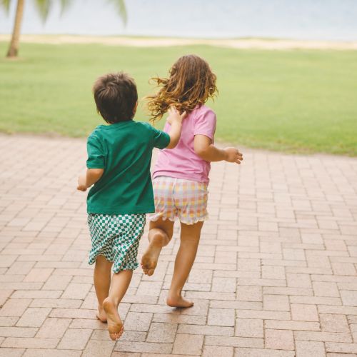 Two children running barefoot on a paved path outdoors, seen from behind. One child wears a green shirt and green-and-white checkered shorts, and the other wears a pink shirt and a light plaid skirt.
