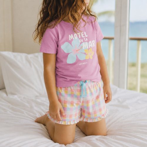 A child kneeling on a white bed wearing a pink short-sleeve pajama T-shirt with a large flower graphic and the words 'Motel de la Mar' and pastel checkered pajama shorts with a light blue drawstring.