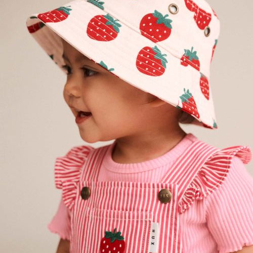 A toddler wearing a white bucket hat with a red strawberry print and a pink striped ruffle-shoulder top, shown in profile against a plain light background.