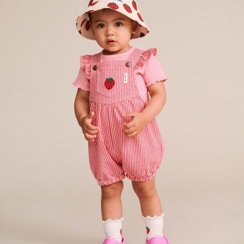 A toddler standing against a plain beige studio background, wearing a pink strawberry-print sun hat, pink and white striped overall romper with ruffled sleeves, white socks, and pink clogs.