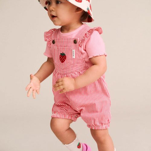 A toddler wearing a pink and white striped short-sleeve romper with ruffled shoulders, a matching strawberry-print sun hat, white socks, and pink clogs, posed against a plain light beige studio background.