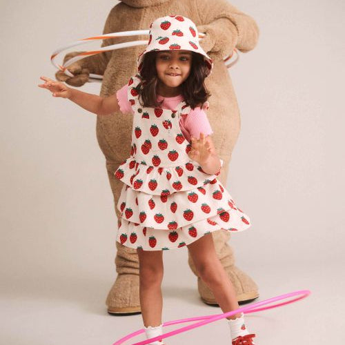 A young girl wearing a strawberry-print dress, matching hat, and red sneakers poses in front of a giant teddy bear while spinning a pink hula hoop on a light beige studio background.