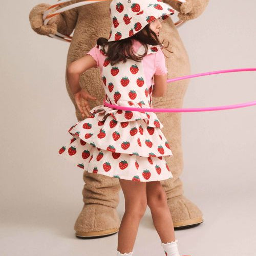 A young girl wearing a strawberry-print tiered dress, matching hat, and red sneakers stands in front of a large teddy bear costume holding hula hoops, photographed against a plain light beige studio background.