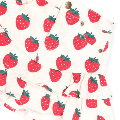 A close-up of a ruffled skirt with a red strawberry print on a cream background, featuring button details and pockets.