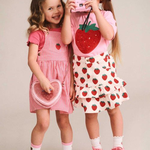 Two young girls modeling strawberry-themed clothing against a plain beige studio background. The girl on the left wears a pink and white striped pinafore dress with a strawberry motif, holding a pink heart-shaped accessory.