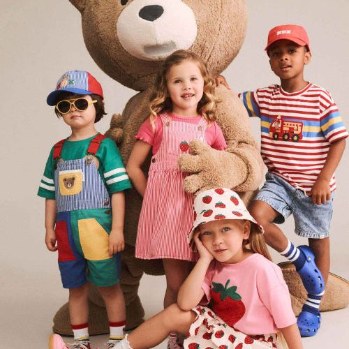 Four children posing with a giant teddy bear in a studio setting. The children are wearing colorful, playful outfits including a pink strawberry-themed dress, a green and blue patchwork overalls set, a red and white striped shirt, and a pink strawberry-patterned hat and skirt.