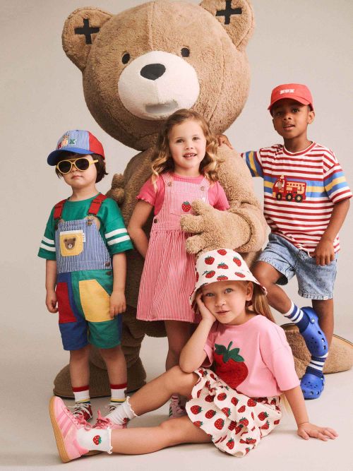 Four children posing with a giant teddy bear in a studio setting. The children are wearing colorful, playful outfits including a pink strawberry-themed dress, a green and blue patchwork overalls set, a red and white striped shirt, and a pink strawberry-patterned hat and skirt.
