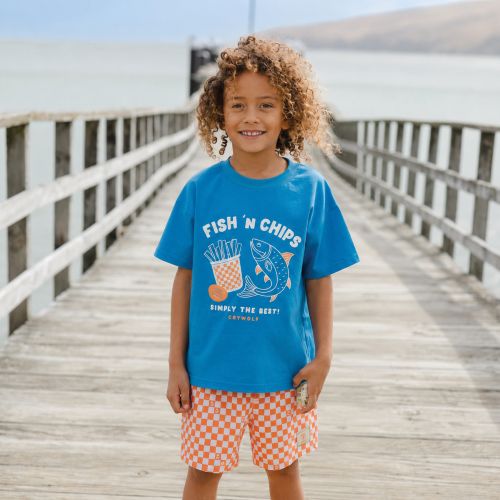 A smiling child standing on a wooden boardwalk by the water, wearing a bright blue graphic T-shirt that reads “FISH ’N CHIPS” with a fish and fries illustration, paired with orange-and-white checkered shorts.