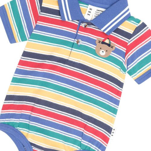 A baby short-sleeve polo romper with horizontal stripes in blue, red, green, yellow, and navy, featuring a small teddy bear patch on the chest and a blue collar.