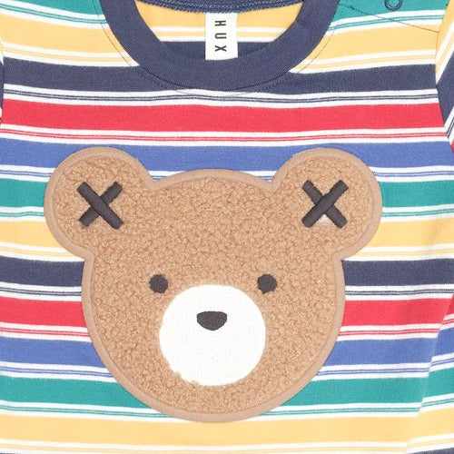 A close-up of a children's striped long-sleeve top with a large brown teddy bear face applique on the front. The shirt features horizontal stripes in navy blue, red, yellow, green, and white, with a navy collar and a white tag reading 'XUX' inside the neck.