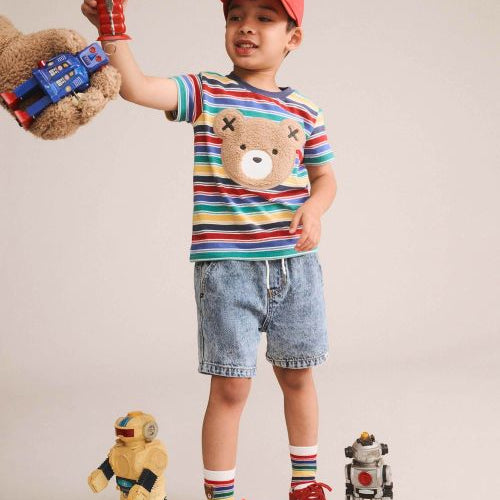 A young boy modeling a colorful striped t-shirt with a teddy bear face graphic, paired with denim shorts, red sneakers, and a red cap. He is holding a toy robot in his raised hand, with small toy robots on the floor against a plain beige studio background.