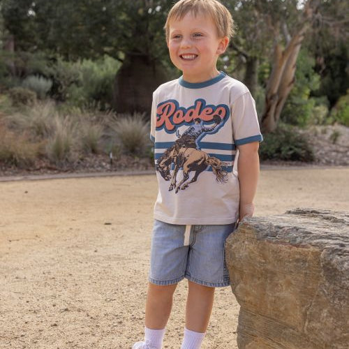 A young boy smiling outdoors next to a large rock, wearing a beige graphic t-shirt with 'Rodeo' and a cowboy print, blue denim shorts, white socks, and white sneakers.