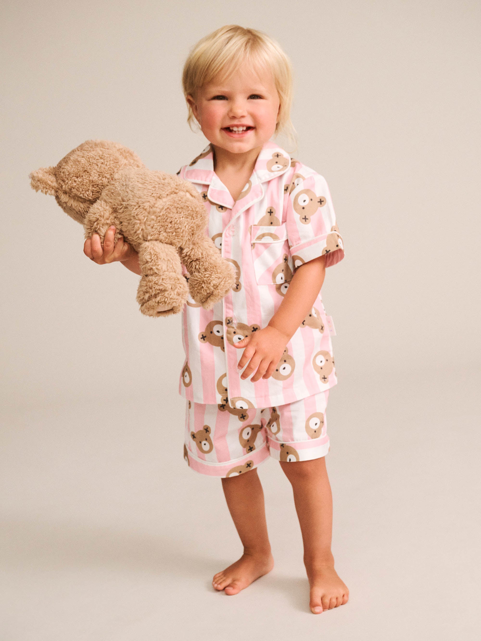 A smiling toddler stands barefoot against a plain light beige studio background, wearing a pink and white striped pajama set with a teddy bear face print and holding a plush teddy bear.