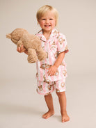 A smiling toddler stands barefoot against a plain light beige studio background, wearing a pink and white striped pajama set with a teddy bear face print and holding a plush teddy bear.