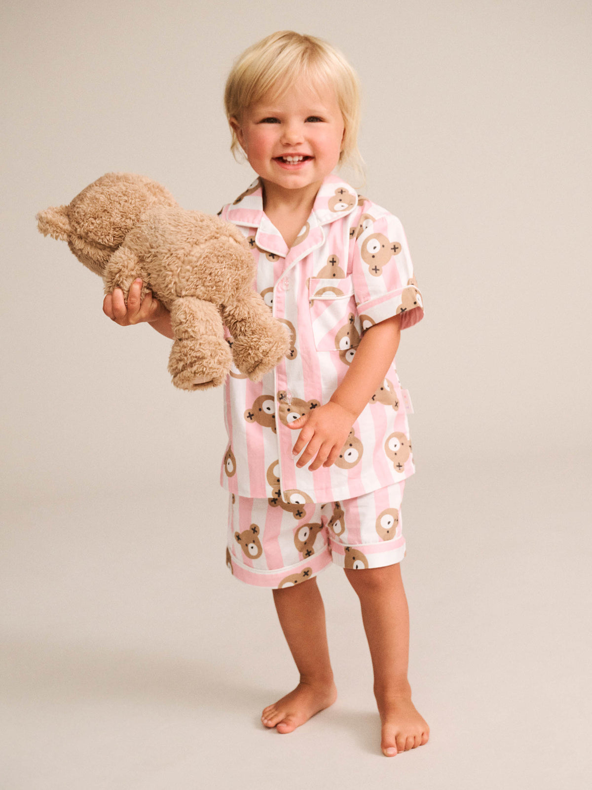A smiling toddler stands barefoot against a plain light beige studio background, wearing a pink and white striped pajama set with a teddy bear face print and holding a plush teddy bear.