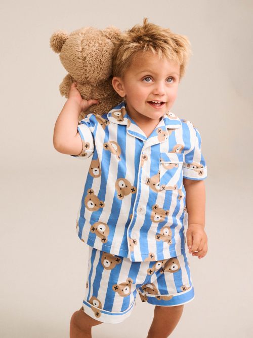 A young child wearing a blue and white striped pajama set with teddy bear prints, holding a brown teddy bear against their back, photographed against a solid light beige background.