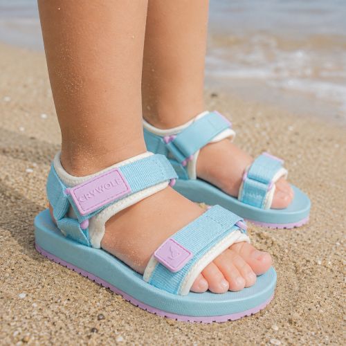 A child wearing light blue and lavender sky-high sandals with white fleece lining, standing on a sandy beach with the ocean in the background.