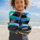 A child holding a pair of Columbia kids' sandals on a beach. The sandals are black with bright blue straps and green accents, including a green brand tab on each strap.