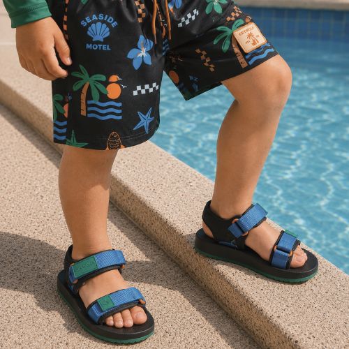 A child wearing black Seaside Motel sandals with blue and green straps and matching black swim shorts with a tropical palm tree print, standing on the edge of a swimming pool.