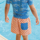 A child standing by a swimming pool, shown from the back wearing a blue short-sleeve swim shirt with a fish print and orange-and-white checkered swim trunks with a blue pocket.