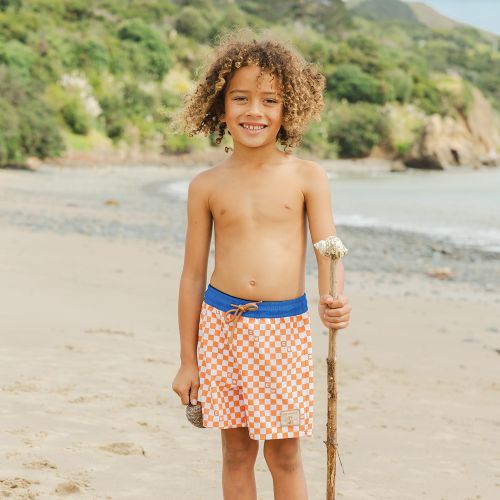 A smiling young boy with curly hair stands on a sandy beach holding a wooden stick, wearing orange and white checkered swim trunks with a blue waistband.