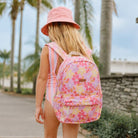 A young girl seen from behind wearing a pink floral backpack, a pink bucket hat, and a pink and white striped swimsuit while walking outdoors on a sunny day with palm trees in the background.