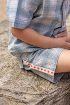 A close-up of a child sitting on a rock, wearing a light blue and white plaid short-sleeve shirt and shorts set with a decorative red star trim on the hem.
