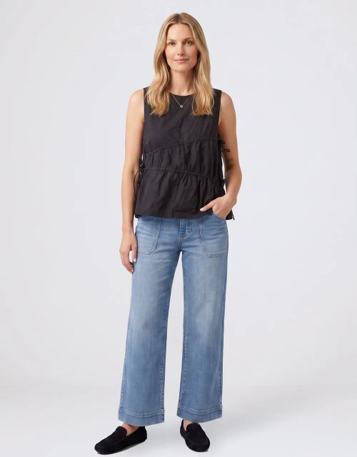 A woman modeling a sleeveless black peplum top with gathered detailing, paired with wide-leg blue jeans and black slip-on shoes, against a light gray studio background.