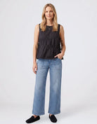 A woman modeling a sleeveless black peplum top with gathered detailing, paired with wide-leg blue jeans and black slip-on shoes, against a light gray studio background.