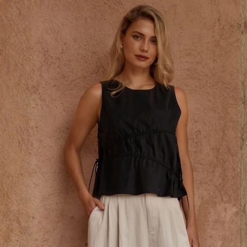 A woman wearing a sleeveless black peplum-style top with gathered detailing and a white skirt, standing against a textured warm beige wall.