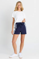 A woman modeling navy blue drawstring shorts with white side stripes, paired with a white t-shirt and white sneakers, against a plain white studio background.