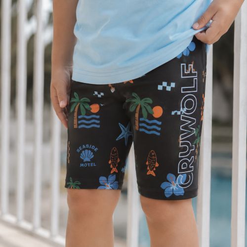 A child wearing black swim trunks with a colorful tropical beach print and the word 'CRYWOLF' printed vertically on the leg, paired with a light blue t-shirt.