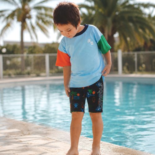 A young boy wearing a light blue, orange, and green color-blocked swim shirt and dark navy swim trunks with a tropical print, standing on the edge of a swimming pool.