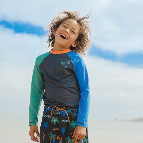A child wearing a long-sleeve rash guard and swim trunks standing on a beach. The rash guard has a dark gray body, green sleeves on the left side, and bright blue sleeves on the right side, with an orange collar and a small palm tree graphic on the chest.